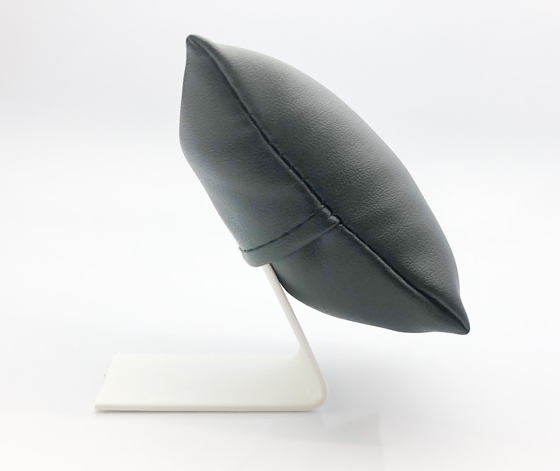 May include: A black leather watch pillow with a white base. The pillow is shaped like a rounded rectangle and is designed to hold a watch.
