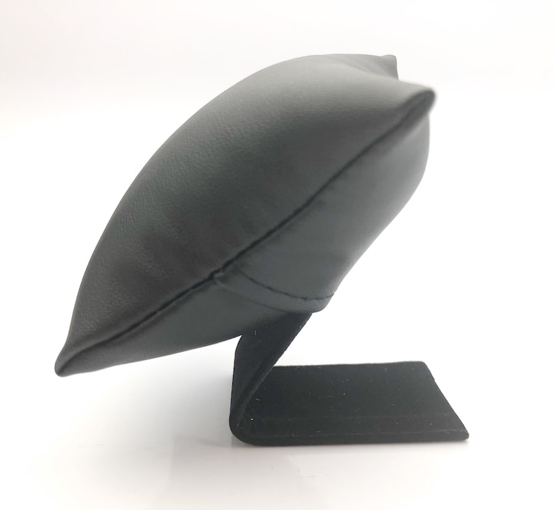 May include: Black leather watch display stand with a pillow-shaped top and a rectangular base.
