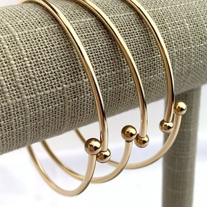 Puede incluir: Tres pulseras de brazalete doradas con un pequeño acento de bola en el extremo de cada pulsera.