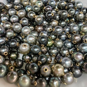 Könnte beinhalten: Eine Ansammlung dunkelgrauer Perlen mit einem glänzenden Schimmer. Jede Perle hat eine goldfarbene Metallkappe mit einem kleinen Loch. Die Perlen variieren in Größe und Farbton, wodurch ein reichhaltiges, strukturiertes Aussehen entsteht. Die Perlen sind wahrscheinlich für die Schmuckherstellung.