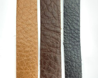 May include: Three strips of textured leather in varying shades of brown and black. The leather has a slightly rough, grainy appearance. The colors include a light tan, a medium brown, and a dark charcoal.
