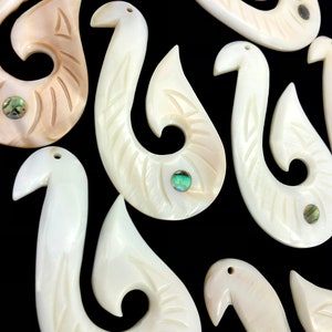 Pode incluir: Um grupo de sete pingentes de anzol de peixe de madrepérola branca esculpida com acentos de concha de abalone verde. Os anzóis são todos do mesmo estilo, com uma forma curva e um pequeno laço na parte superior.