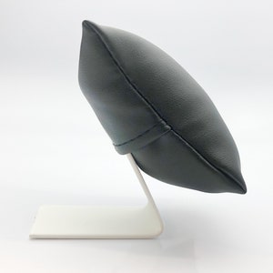 May include: A black leather watch pillow with a white stand. The pillow is shaped like a rounded rectangle and has a seam down the center. The stand is made of metal and has a simple design.
