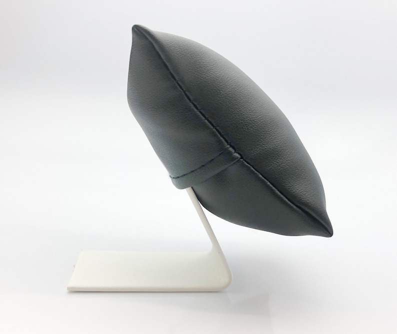 May include: A black leather watch pillow with a white stand. The pillow is shaped like a rounded rectangle and has a seam down the center. The stand is made of metal and has a simple design.