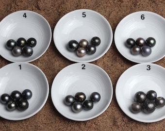 May include: Six white ceramic dishes, each numbered 1 through 6, contain black pearls of varying sizes and shades. The dishes are arranged in two rows of three, on a sandy surface.
