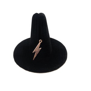 Puede incluir: Un expositor de anillos de terciopelo negro con un dije de rayo dorado en la parte superior.