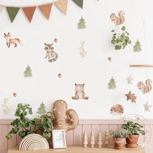 May include: A nursery wall decorated with watercolour illustrations of woodland animals, including a fox, raccoon, bear, rabbit, squirrel, and hedgehog. Green trees and acorns are scattered around the animals. A wooden mobile with stars and a cactus-shaped pillow are also visible.