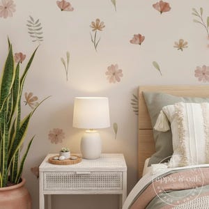 May include: A bedroom scene with a white bedside table, lamp, and floral wall decals. The bed has a decorative pillow and a patterned blanket. A potted snake plant is in the foreground. The lamp is white.