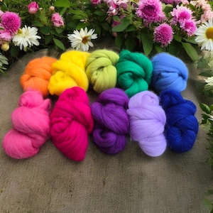 Könnte beinhalten: Eine Sammlung farbenfroher Wollvliese in Rosa, Orange, Gelb, Grün, Blau und Lila. Die Wolle ist in flauschigen, strukturierten Bündeln angeordnet. Im Hintergrund ein Blumenarrangement mit rosa und weißen Blüten.