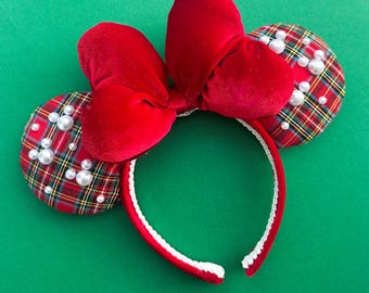 May include: A headband featuring two circular ears covered in a red, green, and yellow plaid fabric adorned with scattered white faux pearls. A large, plush red velvet bow sits between the ears, and the headband band is lined with white trim.