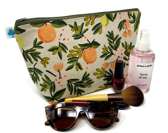 May include: A beige zippered pouch with a floral print featuring green leaves, pink flowers, and orange citrus fruit. The pouch is lying on a white surface with a pair of tortoiseshell sunglasses, a makeup brush, a bottle of nail polish, and a bottle of rose spray.