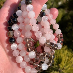 Puede incluir: Tres pulseras de cuentas con cuentas rosas, marrones y negras. Las pulseras están hechas de piedras naturales y están apiladas unas sobre otras.