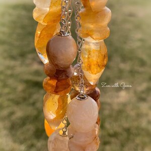 May include: A set of five crystal pendulums with a silver chain. The pendulums are made of clear quartz with inclusions of golden brown and white. The pendulums are hanging from a silver chain. The background is blurred and out of focus. Zen with Gems is written in the bottom right corner of the image.