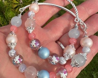 May include: A beaded bracelet featuring Hello Kitty character beads, light blue, pink, and pearl-colored beads. The bracelet has a silver-colored clasp and a twisted silver-colored metal accent.