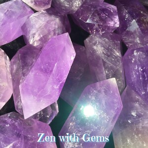 May include: A close-up of a group of amethyst crystals, each with a pointed tip and a smooth, polished surface. The crystals are a deep purple colour and have a slight sheen. The crystals are arranged in a random pattern, with some overlapping others. The text "Zen with Gems" is visible at the bottom of the image.