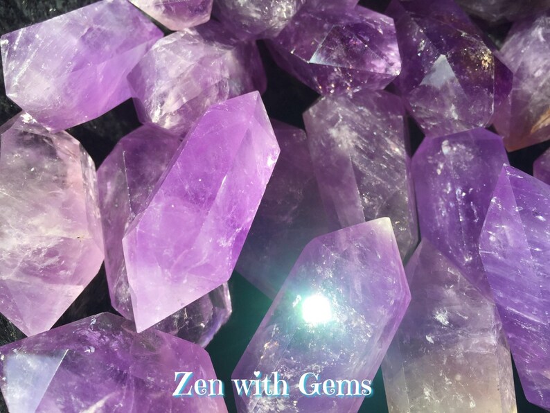 May include: A close-up of a group of amethyst crystals, each with a pointed tip and a smooth, polished surface. The crystals are a deep purple colour and have a slight sheen. The crystals are arranged in a random pattern, with some overlapping others. The text "Zen with Gems" is visible at the bottom of the image.