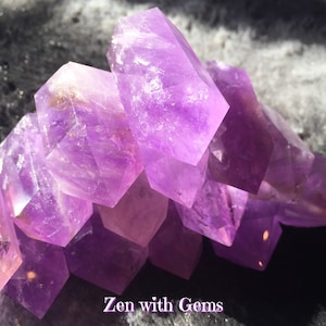 May include: A group of purple amethyst crystals, stacked on top of each other, with a light reflecting off the surface of the crystals. Zen with Gems.