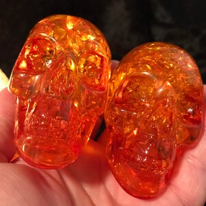 May include: Two amber glass skull figurines. The skulls are detailed and have a translucent, slightly orange hue.