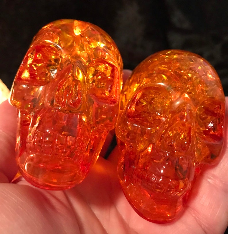 May include: Two amber glass skull figurines. The skulls are detailed and have a translucent, slightly orange hue.