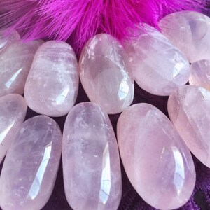 May include: A collection of smooth, polished rose quartz stones in various sizes. The stones are a pale pink color and have a translucent appearance. The stones are arranged on a purple fabric background.