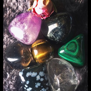 May include: A collection of seven polished gemstones, including amethyst, black obsidian, tiger's eye, malachite, clear quartz, and snowflake obsidian. The stones are arranged on a fuzzy black surface with a dried rose.