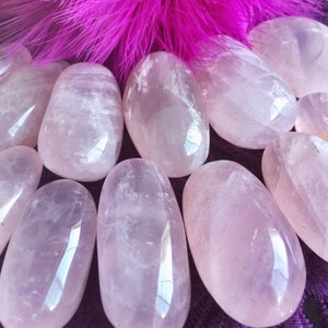 May include: A collection of smooth, oval-shaped rose quartz stones, with a soft pink hue, arranged on a purple velvet surface. The stones are polished and have a translucent appearance.  Zen with Gems is written in white text on a pink heart.