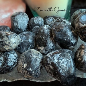 May include: A collection of black obsidian tumbled stones with a white, cloudy, and opaque appearance. The stones are arranged on a white, textured surface.  Zen with Gems.