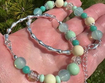 May include: A beaded bracelet with a silver bamboo-style design. The bracelet includes green, cream, and clear beads, and has a silver clasp and extender chain. The bracelet is shown against a natural green backdrop.