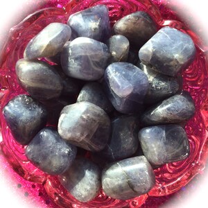 May include: A collection of tumbled blue kyanite stones in a pink bowl. The stones are smooth and have a light blue color with white streaks.