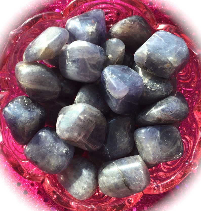 May include: A collection of tumbled blue kyanite stones in a pink bowl. The stones are smooth and have a light blue color with white streaks.