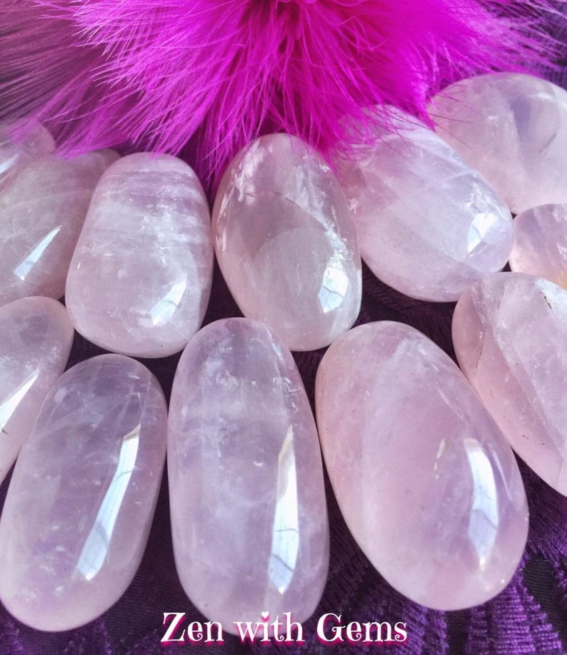 May include: A collection of smooth, polished rose quartz stones in various sizes. The stones are a pale pink color and have a translucent appearance. The stones are arranged on a purple fabric background.