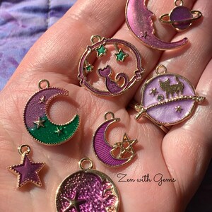 May include: A collection of eight purple and green enamel charms with gold accents. The charms feature a crescent moon, a cat, a star, and a planet. The charms are perfect for adding a touch of whimsy to jewelry or crafts.