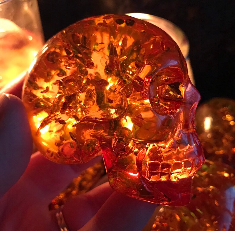 May include: A translucent amber skull sculpture with intricate details. The skull is made of a clear, amber-coloured material and has a detailed, realistic design. The skull is illuminated from behind, creating a warm, glowing effect.