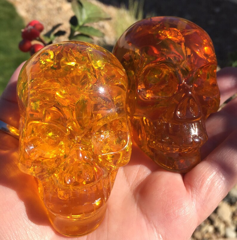 May include: Two amber glass skull sculptures. The skulls are translucent and have a detailed design. The skulls are sitting on a hand.