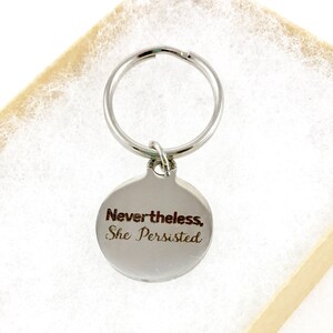 Silver keyring with quote, "Nevertheless, she persisted," in a kraft gift box.