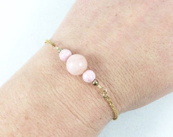 Könnte beinhalten: Ein zartes Armband aus goldener Kette mit drei rosafarbenen Perlen. Die mittlere Perle ist größer als die beiden kleineren Perlen an beiden Seiten.