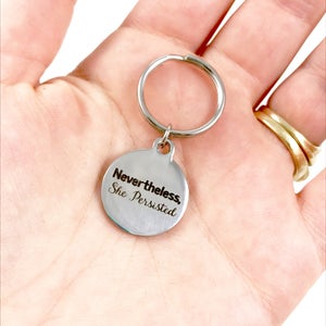 White person's hand holding a silver keyring with quote, "Nevertheless, she persisted."