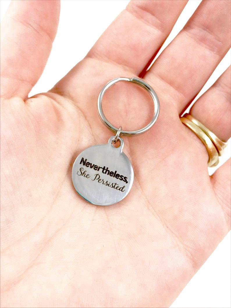 White person's hand holding a silver keyring with quote, "Nevertheless, she persisted."