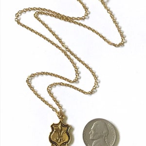 A gold tone brass pendant with fleur de lis design dangles from a 16k gold plated chain with lobster clasp closure with a US nickel coin for size comparison.