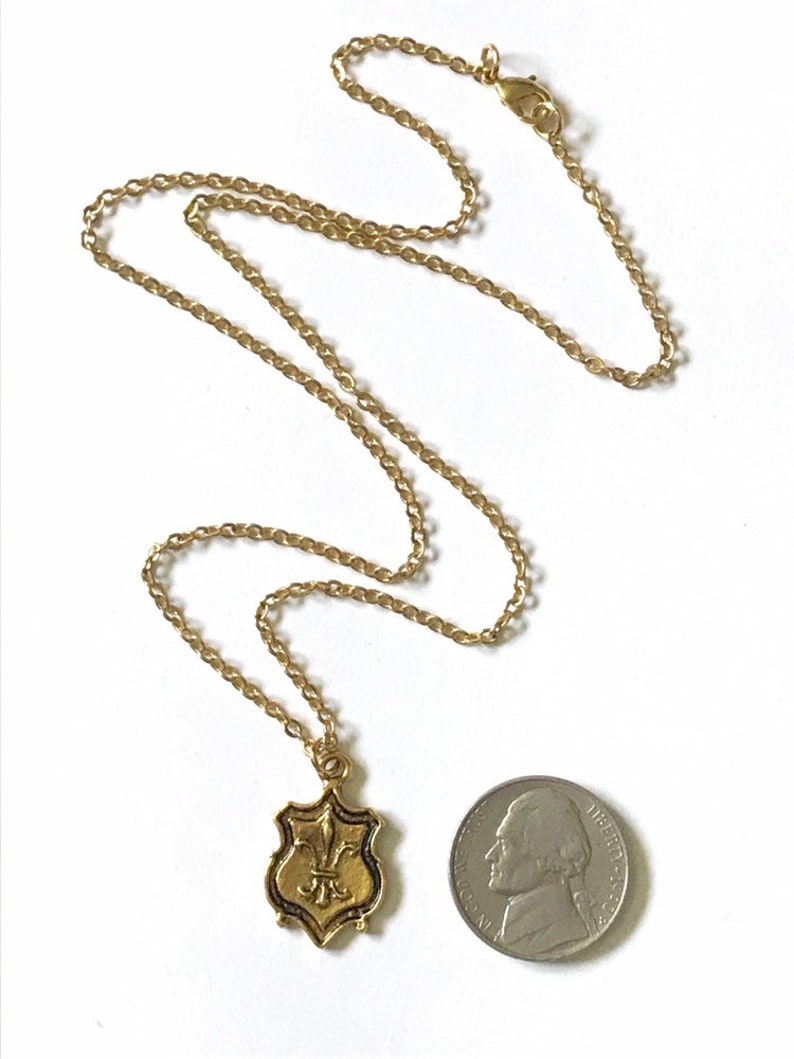 A gold tone brass pendant with fleur de lis design dangles from a 16k gold plated chain with lobster clasp closure with a US nickel coin for size comparison.