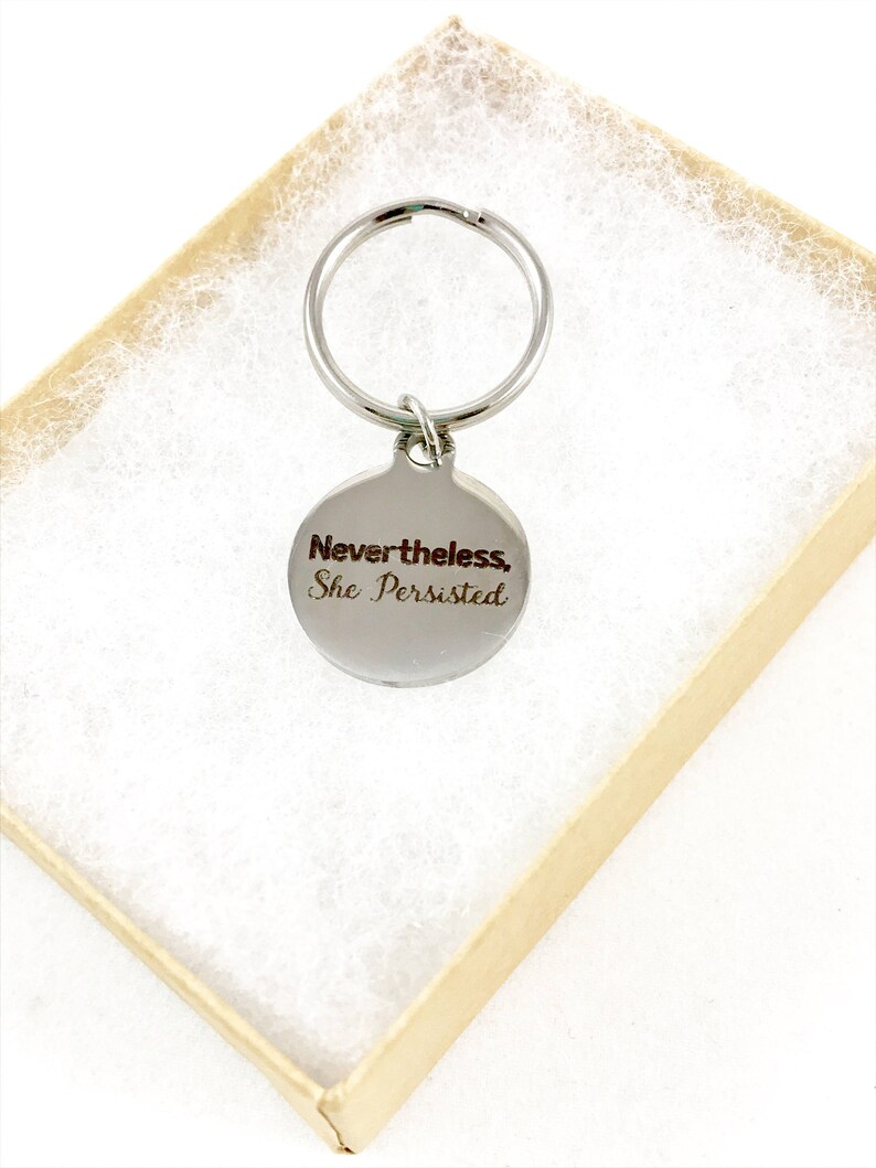 Silver keyring with quote, "Nevertheless, she persisted," in a kraft gift box.