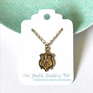 A gold tone brass pendant with fleur de lis design dangles from a 16k gold plated chain. Props include a mint green decorative plate and white jewelry card that says The Joyful Jewelry Box.