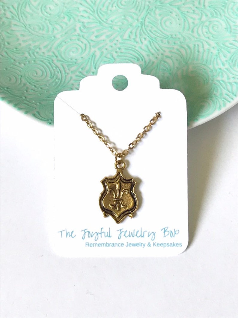 A gold tone brass pendant with fleur de lis design dangles from a 16k gold plated chain. Props include a mint green decorative plate and white jewelry card that says The Joyful Jewelry Box.