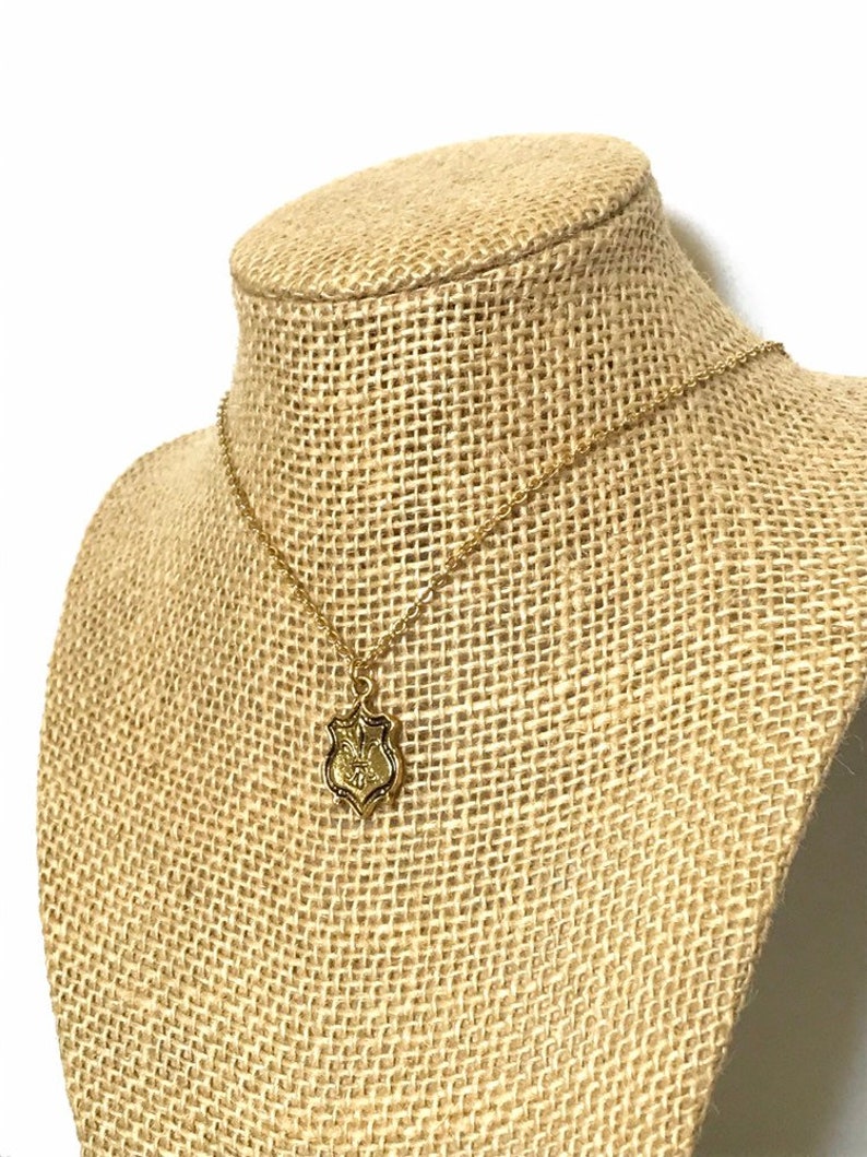 A gold tone brass pendant with fleur de lis design dangles from a 16k gold plated chain with lobster clasp closure. Necklace is displayed on a burlap neck form.