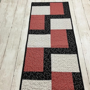 May include: A rectangular quilted table runner with a patchwork design. The runner features alternating red and white patterned squares, framed by black fabric with a white swirl pattern. The runner is placed on a light wood surface.