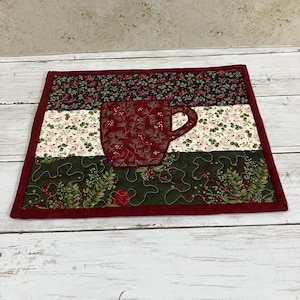 May include: A rectangular quilted fabric mat featuring a central dark red mug appliqué with a pine needle pattern. The mat is divided into three horizontal sections with floral and berry prints in dark green, cream, and black, finished with a red border.