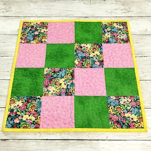May include: A square quilt with a patchwork design. The quilt features alternating squares of pink, green, and floral fabric. The floral fabric has a black background with yellow, pink, and blue flowers. The quilt is bordered with a yellow fabric.