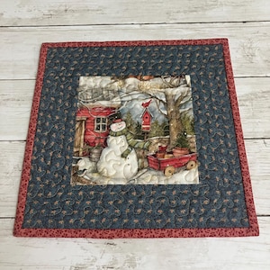 May include: A quilted placemat featuring a winter scene with a snowman, red birdhouse, and a red wagon. The central image is framed by a blue fabric with a floral pattern, and a red border. The placemat is square.