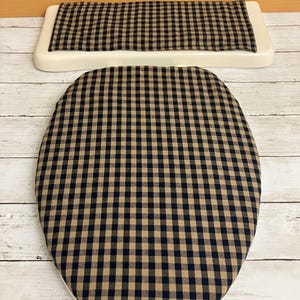 May include: A white toilet seat and lid set with a black and tan gingham pattern. The seat is oval-shaped, and the lid is rectangular. The fabric has a classic, country-style design.
