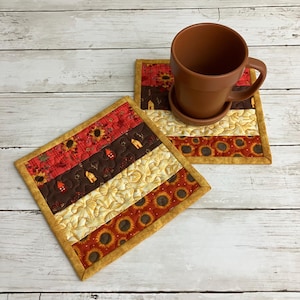 May include: Two square quilted fabric coasters featuring horizontal stripes of sunflower, house, and leaf patterns in shades of red, brown, yellow, and orange. A solid brown ceramic mug with a matching saucer sits on one of the coasters.
